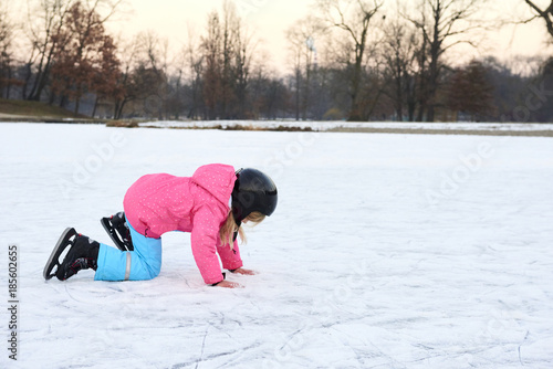 Child girl falling down on ice in snowy park during winter holidays. Wearing safety helmet. Winter children activities.