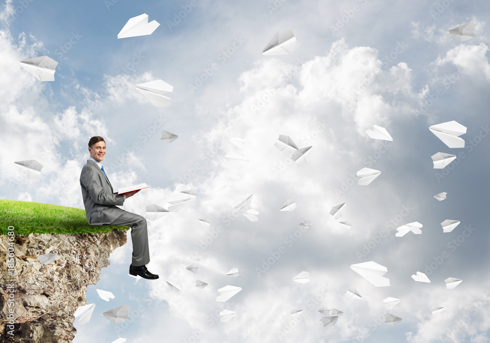 Man on edge reading book and paper planes flying in air Man on edge reading book and paper planes flying in air