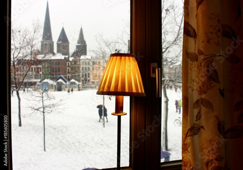 Fototapeta Naklejka Na Ścianę i Meble -  Winter square with snow view from window. Cozy interior with yellow lamp shade and curtains. Old buildings, castle and church landmark in Europe. Winter european town landscape.