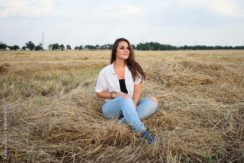 attractive smiling brunette in white shirt and jeans sitting on haystack in front of field. attractive smiling brunette in white shirt and jeans sitting on haystack in front of field.