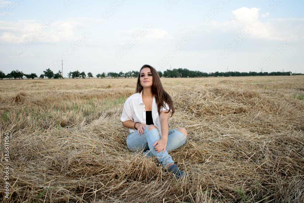 attractive smiling brunette in white shirt and jeans sitting on haystack in front of field. attractive smiling brunette in white shirt and jeans sitting on haystack in front of field.