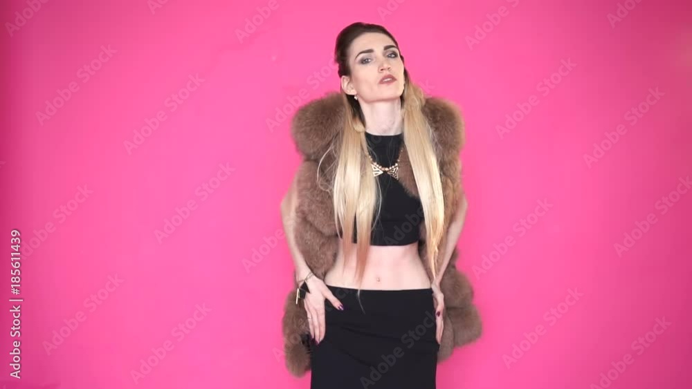 Glamorous caucasian young woman in a stylish fur coat dancing and looking at the camera, isolated on pink background.