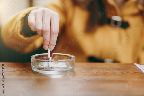 Focus on caucasian young woman hand putting out cigarette on glass ashtray on wooden table, cigarette butt, smoking is dying. Quit smoking. Health concept. Close up photo.