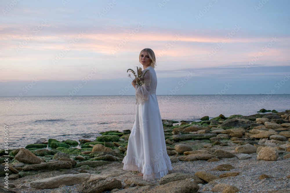 Elegant young woman in white long dress on sea background. Bride with flowers at sunset, sunrise,dawn Elegant young woman in white long dress on sea background. Bride with flowers at sunset, sunrise,dawn