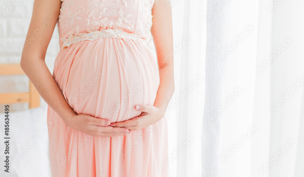 Pregnancy woman in beautiful pink dress standing close up a window in her bedroom. Pregnancy woman in beautiful pink dress standing close up a window in her bedroom.