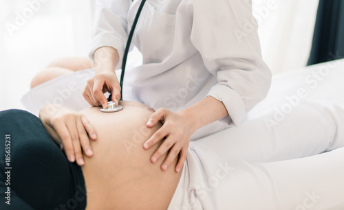 Female Obstetrician doctor with stethoscope listening to pregnant woman baby heartbeat at hospital.