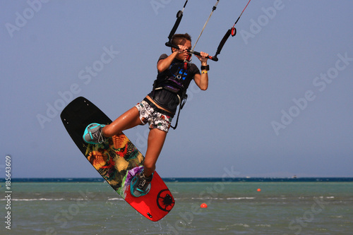 Kitesurfing girl in boots and shorts with kite in blue sky on kiteboard in the blue sea jumping air trick. Recreational activity, water sports, action, hobby and fun in summer time. Kiteboarding sport