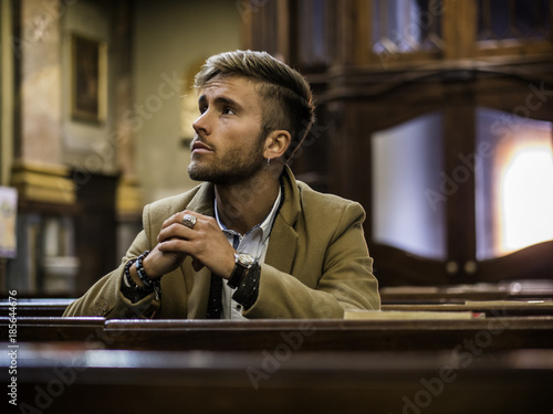 Young blond elegant man sitting in church, on wooden bench, thinking and praying, looking around