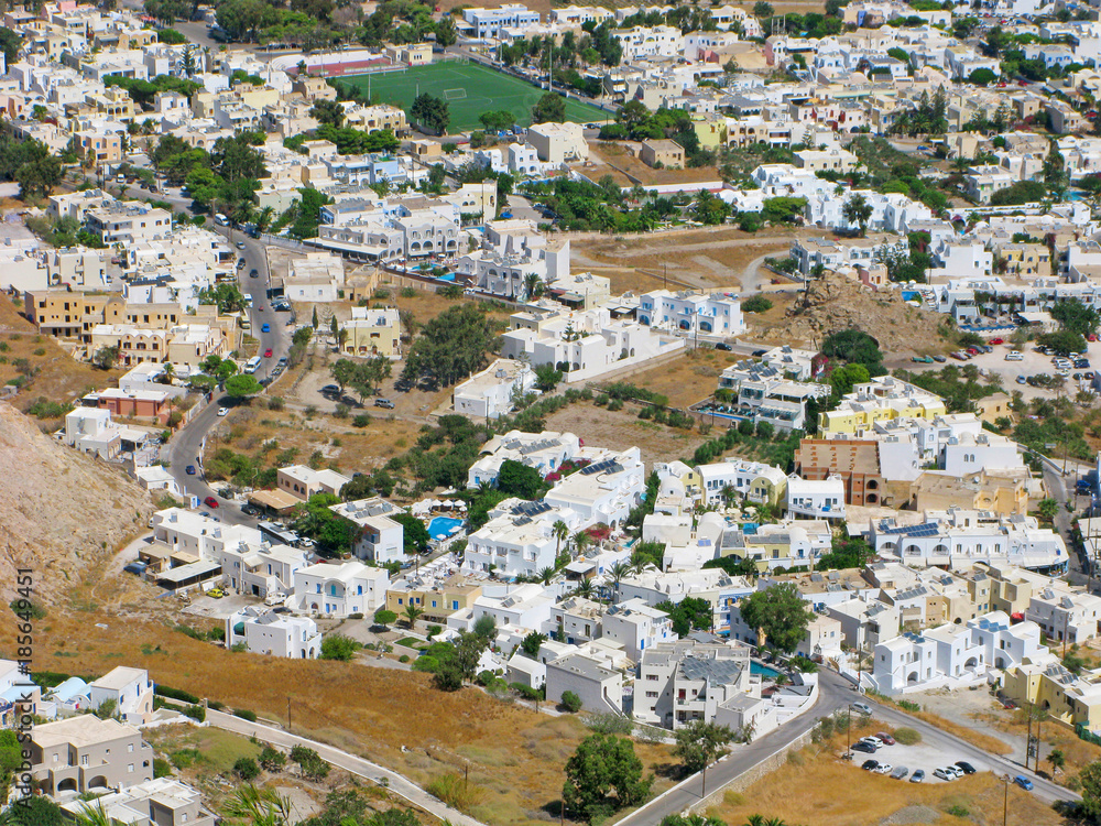 Cyclades, Greece. Kamari village on Santorini from aerial view. Low-rise houses, cars on road, green stadium for sport. Urban skyline Cycladic architecture.