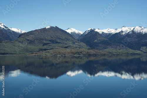 Paisaje de montañas frente a un gran lago donde se reflejan. Cielo azul despejado.