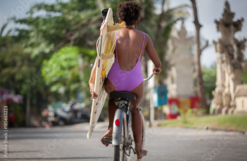Beautiful surfer girl in purple bikini with afro hairstyle riding bicycle with one hand, carrying surfboard under her arm at Kuta beach, Bali, Indonesia © Hagen Production