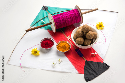 happy Makar Sankranti Festival - Tilgul or Til ladoo in a bowl or plate with haldi kumkum and flowers with Fikri /Reel/Chakri /Spool with colourful thread or manjha and kite over plain background