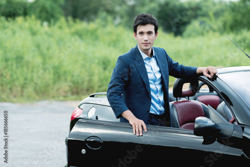 Holiday Car rent and road trip concept. Handsome white man standing on a side of black convertible with nature background, looking into camera, ready for a road trip and long drive.
