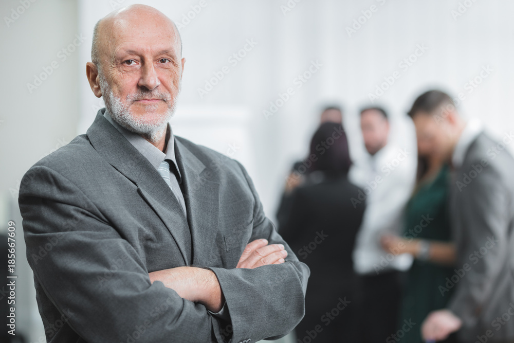 Portrait of serious senior businessman standing with arms folded in office lobby, colleagues in background.