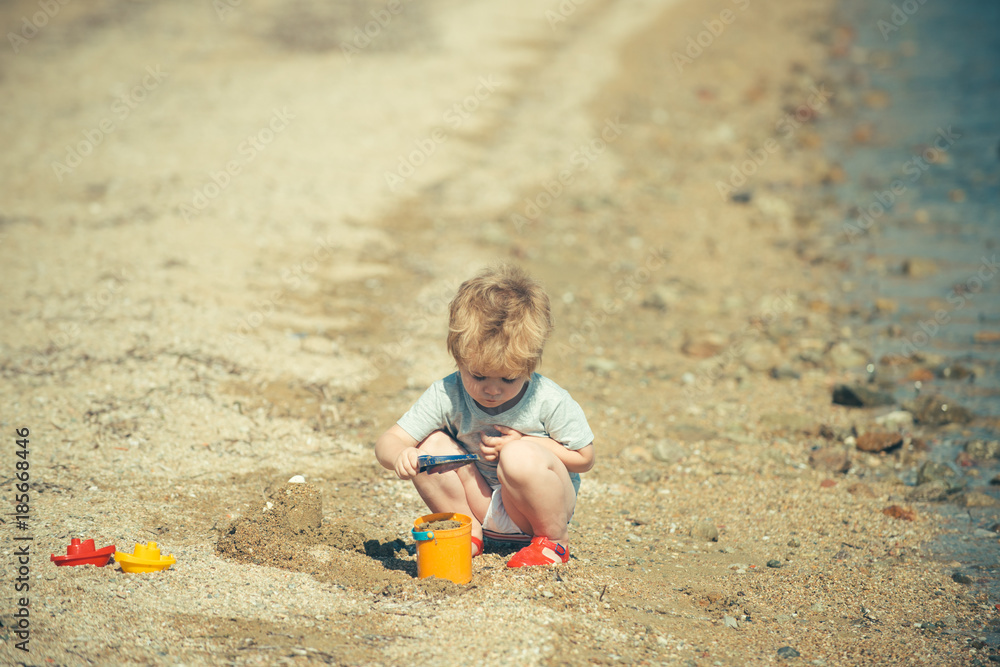 Beach game with sand. Cute boy is playing near sea or ocean with bucket and shovel, builds sand castles. Childhood and happy summer, seaside holidays Beach game with sand. Cute boy is playing near sea or ocean with bucket and shovel, builds sand castles. Childhood and happy summer, seaside holidays