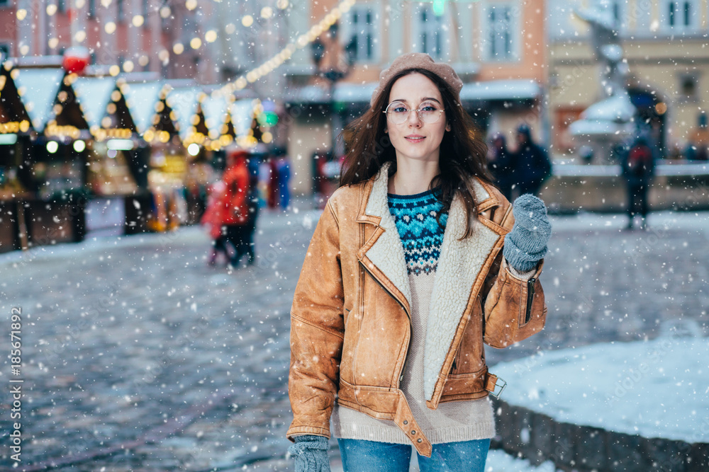 Portrait of woman in woolen sweater, beret and leather jacket enjoying winter moments. Outdoor photo of long-haired inspiring lady having fun in snowy evening over old town square with Christmas fair.