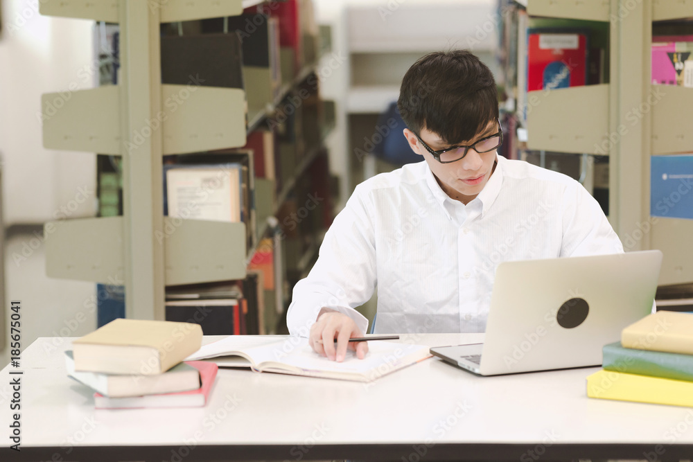 Young student study hard in library. Asian male university student doing study research in library with books and laptop on desk and smiling. For back to school education technology concept. Young student study hard in library. Asian male university student doing study research in library with books and laptop on desk and smiling. For back to school education technology concept.
