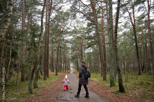 Little girl wear jacket and hat walk is playing at the forest with mother near baltic sea on cloudy sky in winter time. back view