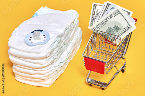 babies diapers with pacifier near shopping trolley cart with money and baby stuff on yellow background