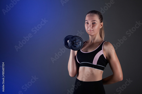 A sporty girl holds a dumbbell in her hands, shakes a muscular. Against a dark background