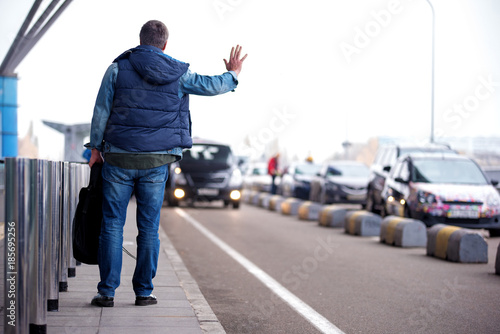 Photography Full length back view of adult man with backpack is standing near road while raising hand