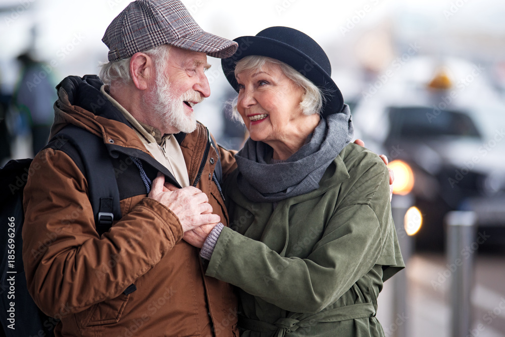 My love with you. Waist up of pleasant elderly bearded man with backpack and charming old woman are standing outdoors and hugging. They are looking at each other with fondness