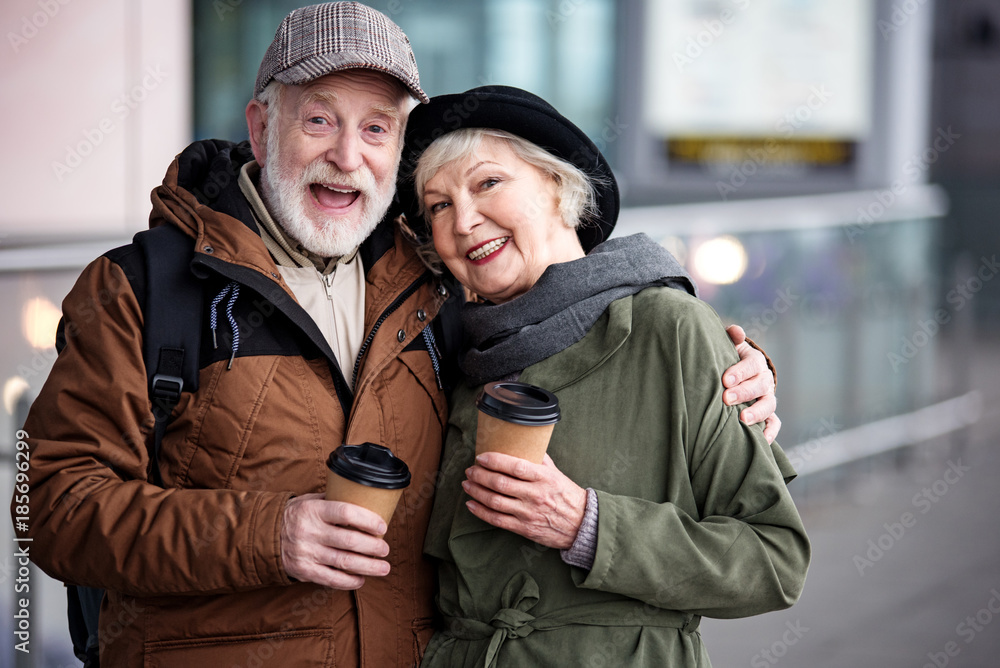 Together forever. Waist up portrait of happy aged couple is standing together outdoors and holding coffee. Men is hugging elegant woman and expressing admiration. They are looking at camera with joy Together forever. Waist up portrait of happy aged couple is standing together outdoors and holding coffee. Men is hugging elegant woman and expressing admiration. They are looking at camera with joy