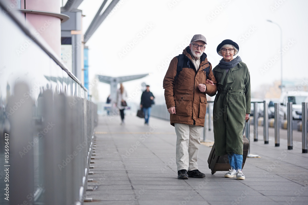 Obraz premium My faithful companion. Full length portrait of aged couple is standing with their luggage outdoors near international airport. They are looking at camera with smile. Copy space in the left side