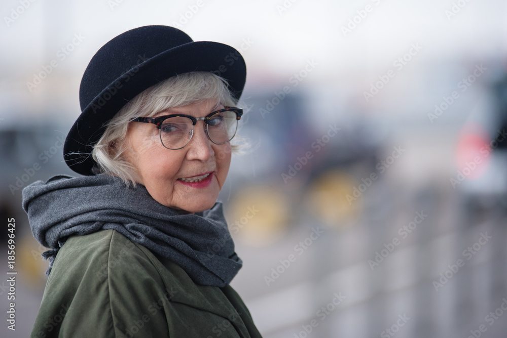 Pleasant lady. Portrait of charming stylish senior female in glasses is standing outside and looking at camera with slight smile. Copy space in the right side Pleasant lady. Portrait of charming stylish senior female in glasses is standing outside and looking at camera with slight smile. Copy space in the right side