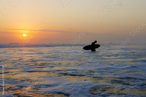 Silhouette of unknown anonymous surfer holding surf board after surfing on sunset with amazing beautiful sunlight with orange sky