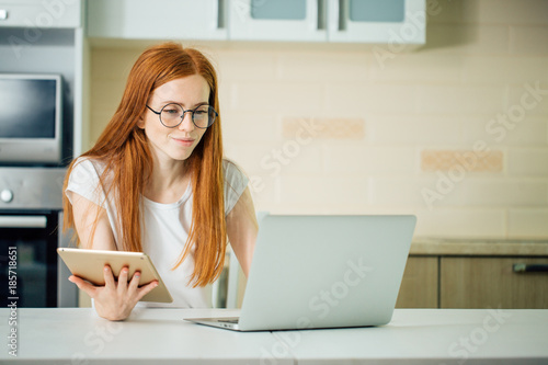 young redhead woman using the tablet pc and laptop in kitchen photo