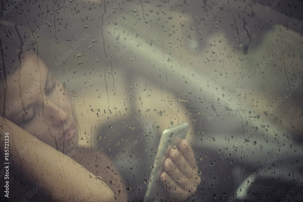Sad woman looking in the mobile phone and reading message in the window with rain drop in the car. Face of young female behind rain car window. Loneliness and depression concept. Psychology Sad woman looking in the mobile phone and reading message in the window with rain drop in the car. Face of young female behind rain car window. Loneliness and depression concept. Psychology