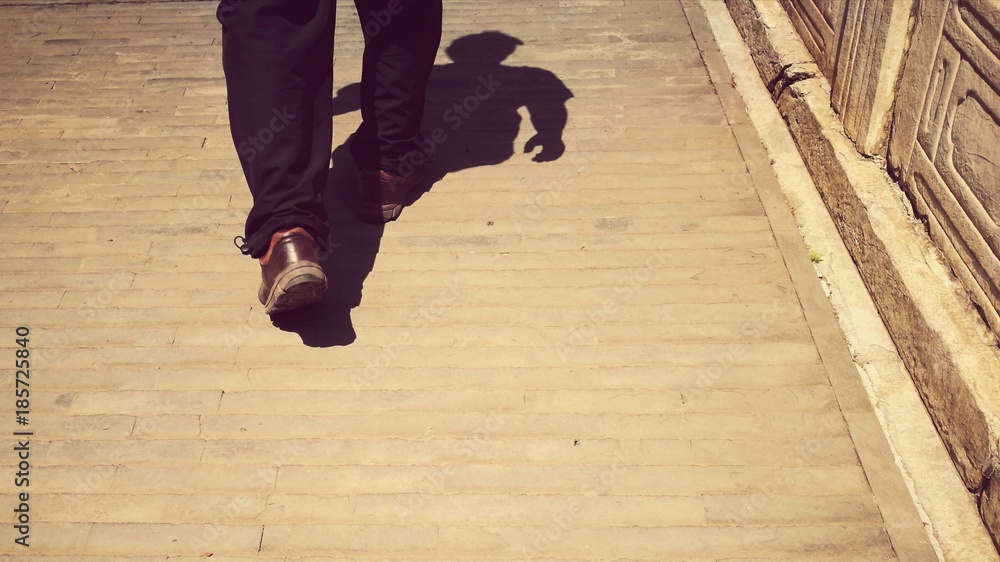 Business man walking on an ancient city sidewalk (in China). The image was taken from waist down with shadow of the whole body; retro style; bottom copy space for text Business man walking on an ancient city sidewalk (in China). The image was taken from waist down with shadow of the whole body; retro style; bottom copy space for text