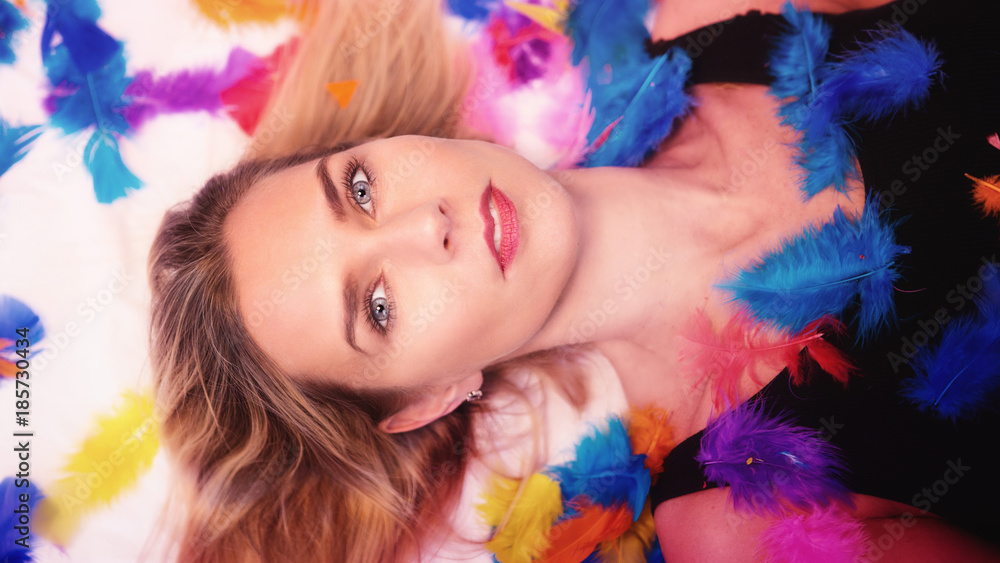 blond woman lying down surrounded by colorful feathers blond woman lying down surrounded by colorful feathers