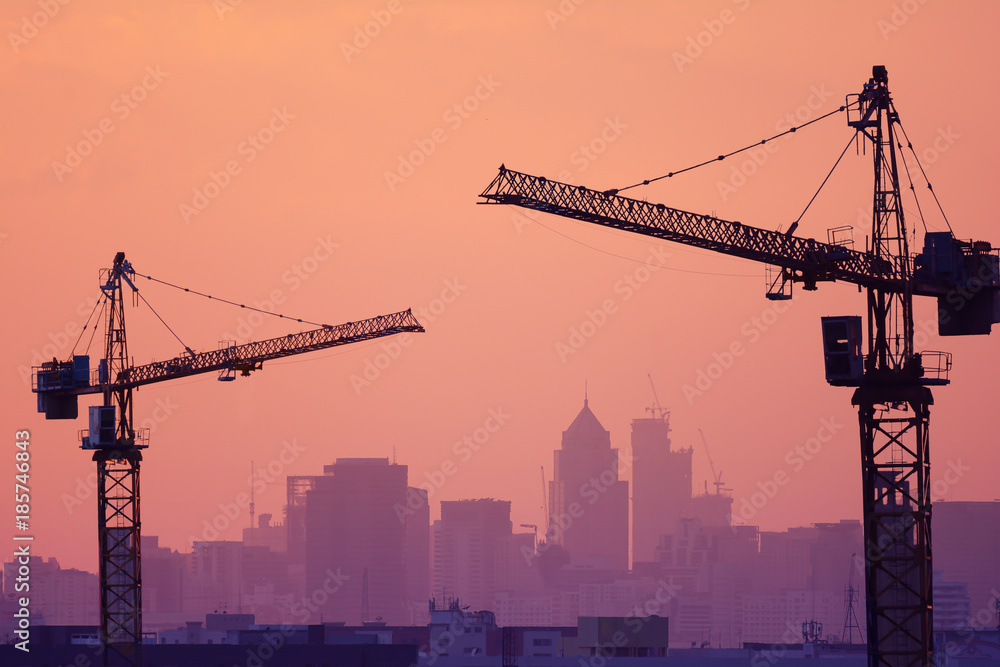 Construction site have two cranes and purple silhouettes of building and orange background.