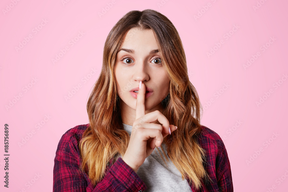 Beautiful young female with appealing appearance keeps fore finger on lips, demonstrates silence sign, asks to be quiet, isolated over pink studio background. Hush gesture. Woman gestures indoor Beautiful young female with appealing appearance keeps fore finger on lips, demonstrates silence sign, asks to be quiet, isolated over pink studio background. Hush gesture. Woman gestures indoor