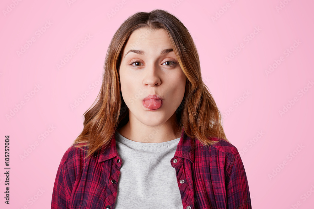 Funny pretty woman foolishes indoor, shows tongue, makes grimace, wears checkered chemise, isolated over pink studio background. Cute young female has fun, demonstrates tongue at camera indoor Funny pretty woman foolishes indoor, shows tongue, makes grimace, wears checkered chemise, isolated over pink studio background. Cute young female has fun, demonstrates tongue at camera indoor