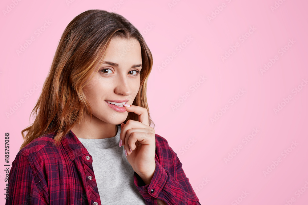 Portrait of pretty woman stands sideways, has delightful look, keeps finger on lips, looks confidently into camera, isolated over pink background with copy space for your advertising content Portrait of pretty woman stands sideways, has delightful look, keeps finger on lips, looks confidently into camera, isolated over pink background with copy space for your advertising content