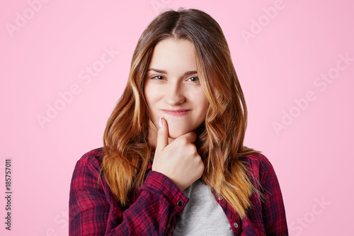 Portrait of attractive woman with intriguing expression keeps hand under chin, curves lips, intends to do something forbidden, has pleased expression, isolated over pink background. Facial expressions