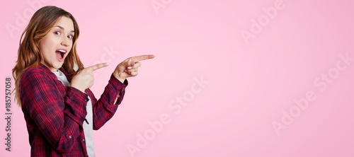 Fotografía Excited beautiful woman stands sideways, wears chemise, points with fore fingers aside at blank copy space for your advertisment or promotional text
