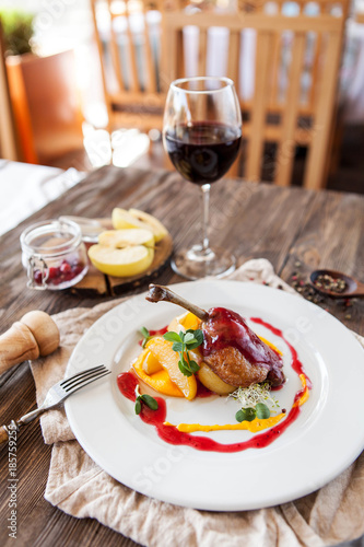 baked duck leg with berry sauce with apples on a white plate with a glass of red wine on a wooden table