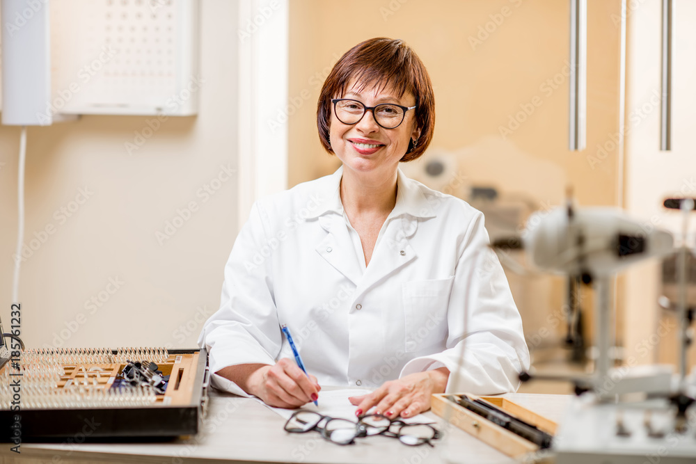 Portrait of a senior woman ophthalmologist in unifrom sitting with different optical devices and eyeglasses during the work in the office