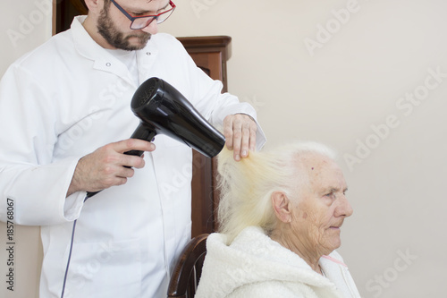 Pielęgniarz suszy suszarką włosy starej kobiety siedzącej na krześle.