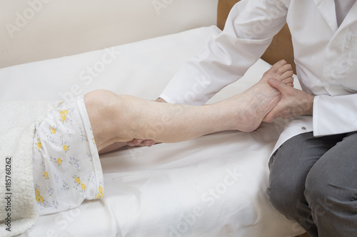 Masażysta masuje łydkę starej kobiety leżącej na łóżku na białym prześcieradle.