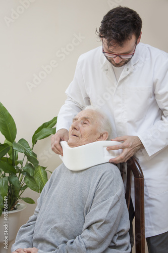 Lekarz ortopeda zakłada biały kołnierz ortopedyczny bardzo starej kobiecie siedzącej na krześle.