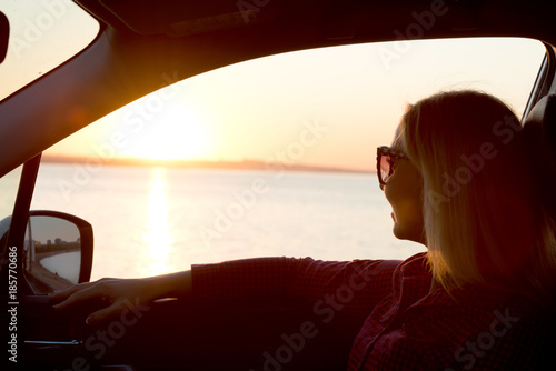 A young woman looks out the car window at the sunset on the sea.	