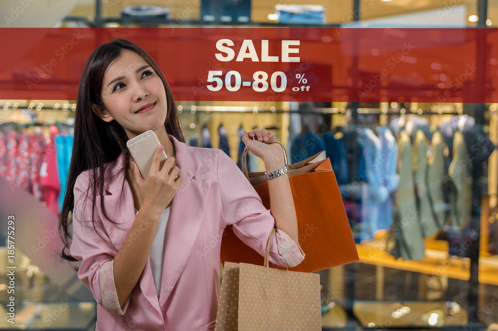 Happy young asian woman shopping in happy feeling and holding the product paper bag over the Sale 50-80% off mock up advertise display frame setting over the Abstract blurred photo of clothing store Happy young asian woman shopping in happy feeling and holding the product paper bag over the Sale 50-80% off mock up advertise display frame setting over the Abstract blurred photo of clothing store