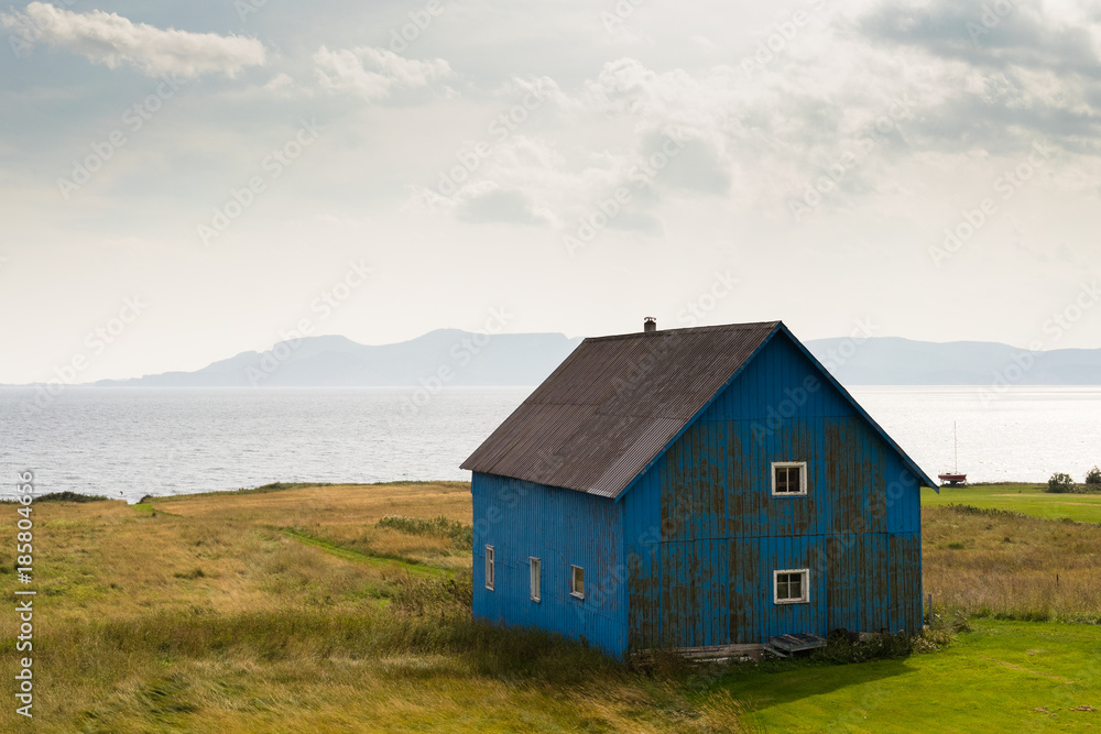 A blue wooden farmhouse with white window frames  standing alone in a field in front of a bay of water along the coastline.