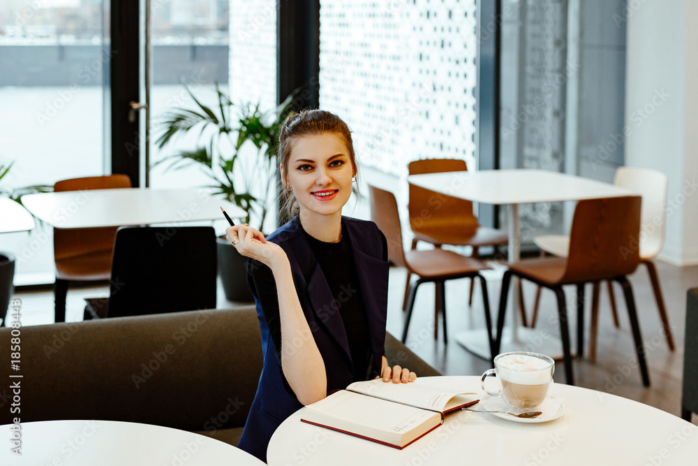 The young business woman in a strict suit makes entries in a notebook. The girl sits in cafe of office and the cup of coffee stands nearby. The young business woman in a strict suit makes entries in a notebook. The girl sits in cafe of office and the cup of coffee stands nearby.