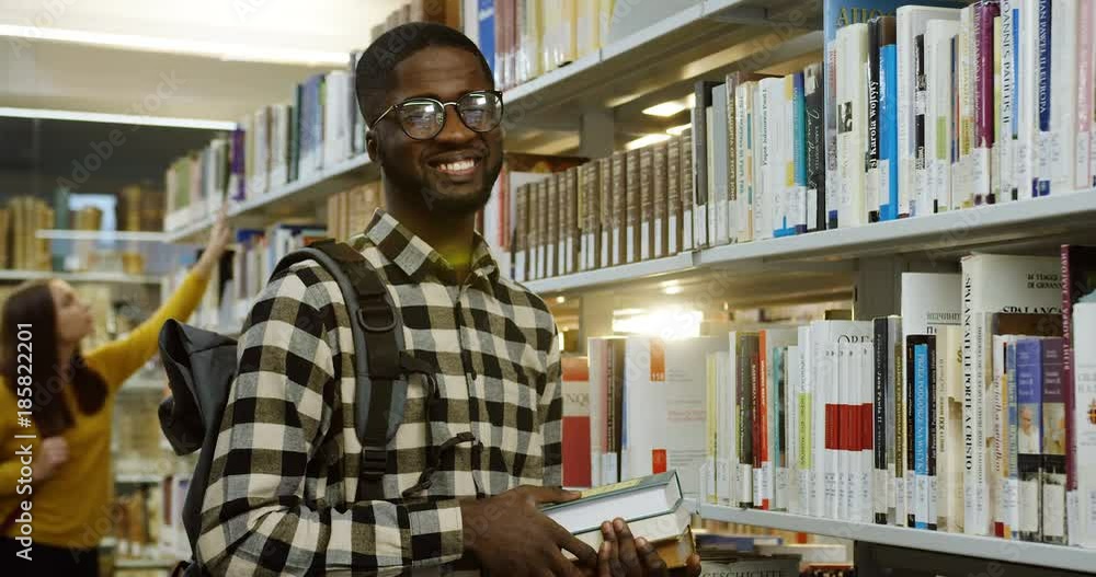 Portrait of the young male African American student in glasses posing and smiling to the camera with books in hands at the library passage with other student on the background. Indoor Portrait of the young male African American student in glasses posing and smiling to the camera with books in hands at the library passage with other student on the background. Indoor